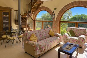 Un amplio salón con sofá, sillón y una mesita de centro. Se observa una escalera con barandillas de madera y vistas al exterior con zonas verdes.