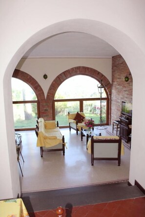 Amplia sala de estar con un gran arco que da a un jardín, un televisor y un comedor con mesa y sillas.