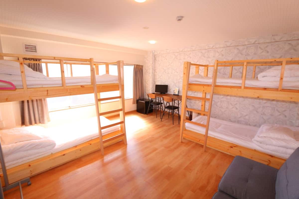 A dormitory room with bunk beds, a desk with a computer, and a small seating area.