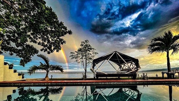 Una estructura moderna con diseño geométrico, reflejada en una piscina cristalina, rodeada de palmeras y una vista al atardecer.