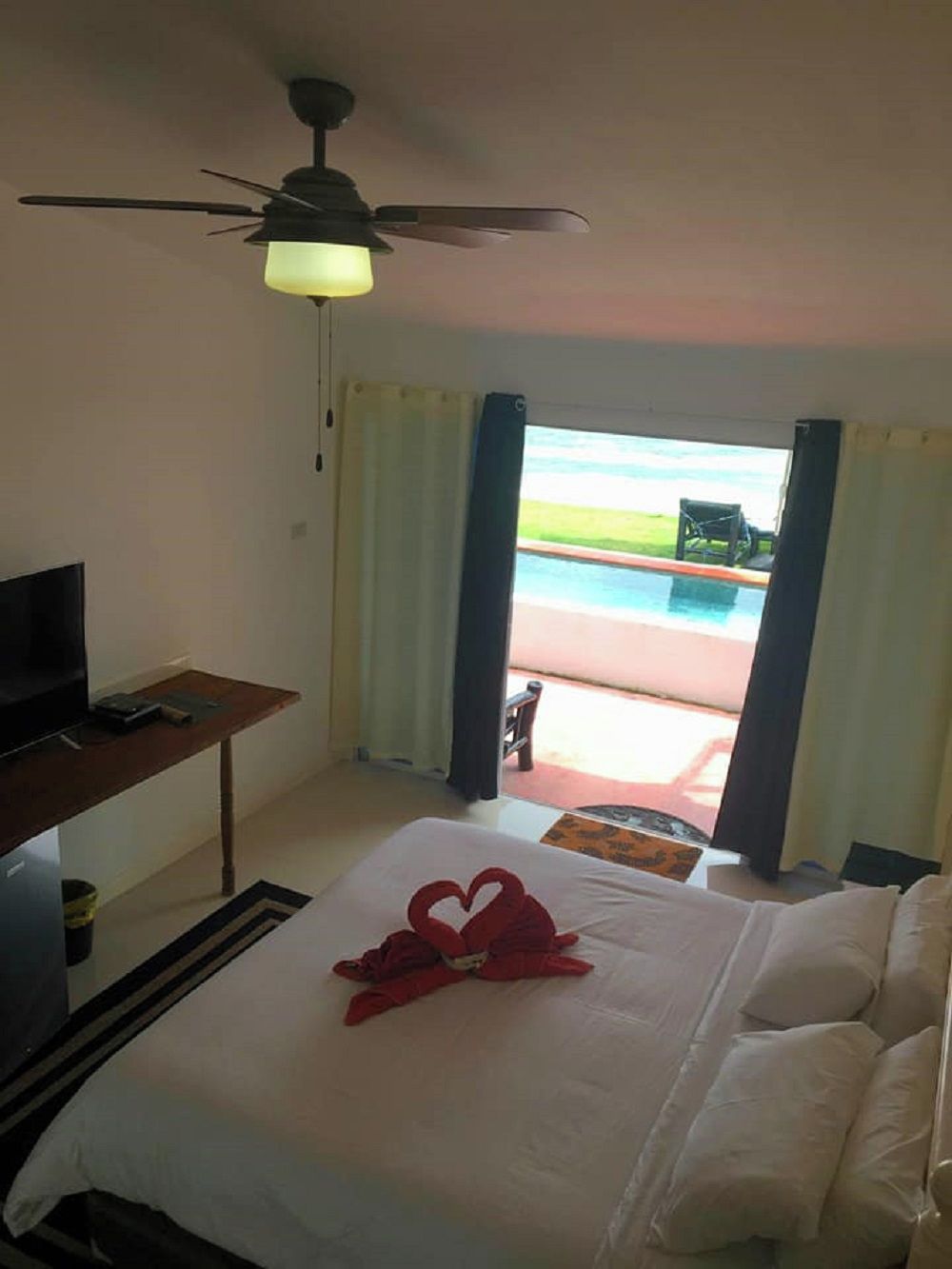 A hotel room with a bed, a television, a ceiling fan, and a view of an outdoor swimming pool.