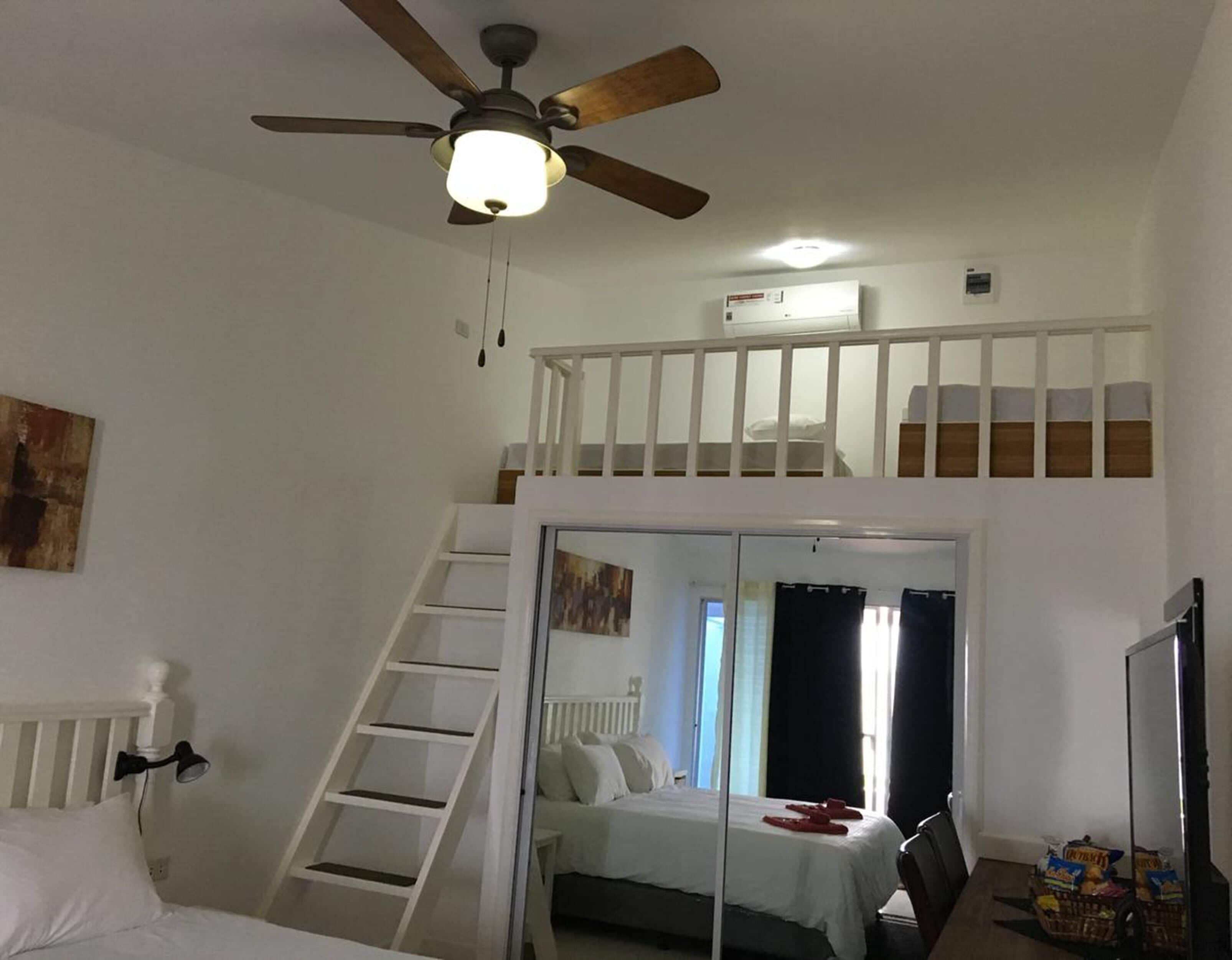 Une chambre avec un lit superposé, un ventilateur de plafond, une échelle, un lit avec une literie blanche, une fenêtre avec des rideaux et un petit coin cuisine équipé d’un micro-ondes et d’un panier de collations.