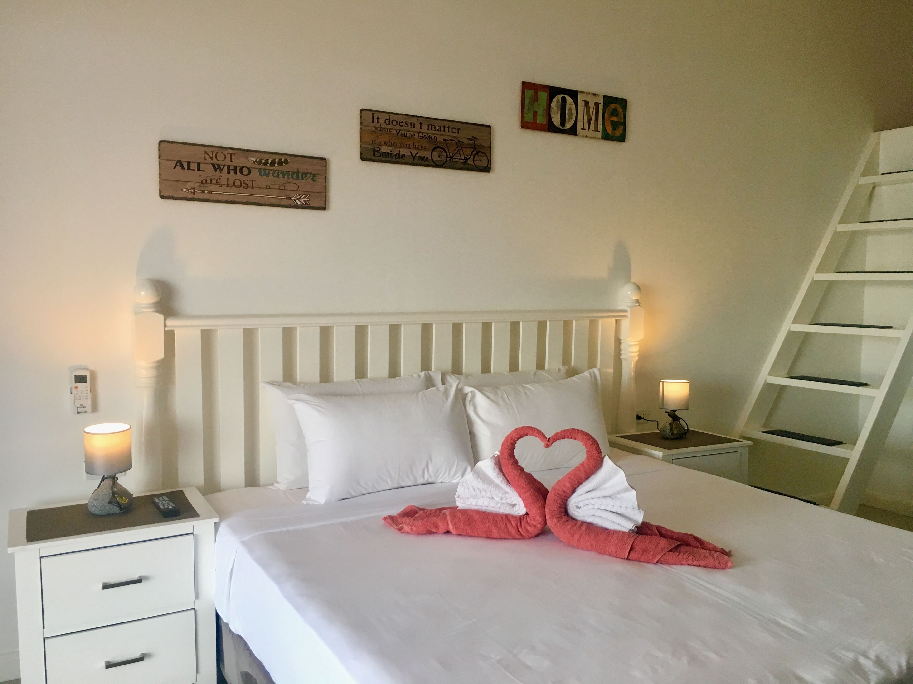 A neatly made bed with white linens and pink towels arranged in a heart shape. There are two bedside tables with lamps, a ladder against the wall, and three motivational signs on the wall.