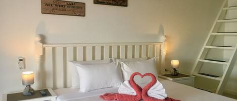 A neatly made bed with white linens and pink towels arranged in a heart shape. There are two bedside tables with lamps, a ladder against the wall, and three motivational signs on the wall.