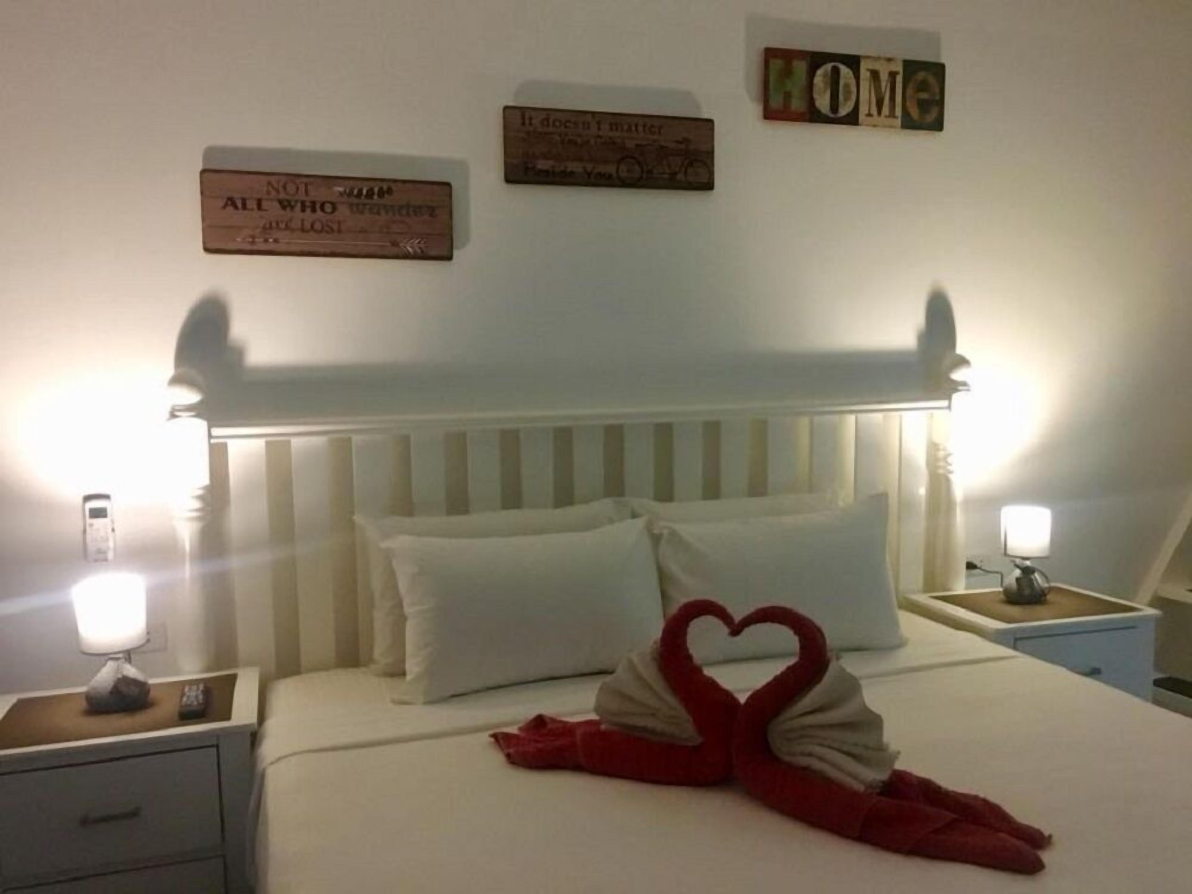 A hotel room with a bed, pillows, a heart-shaped towel arrangement, bedside tables, and wall decorations.