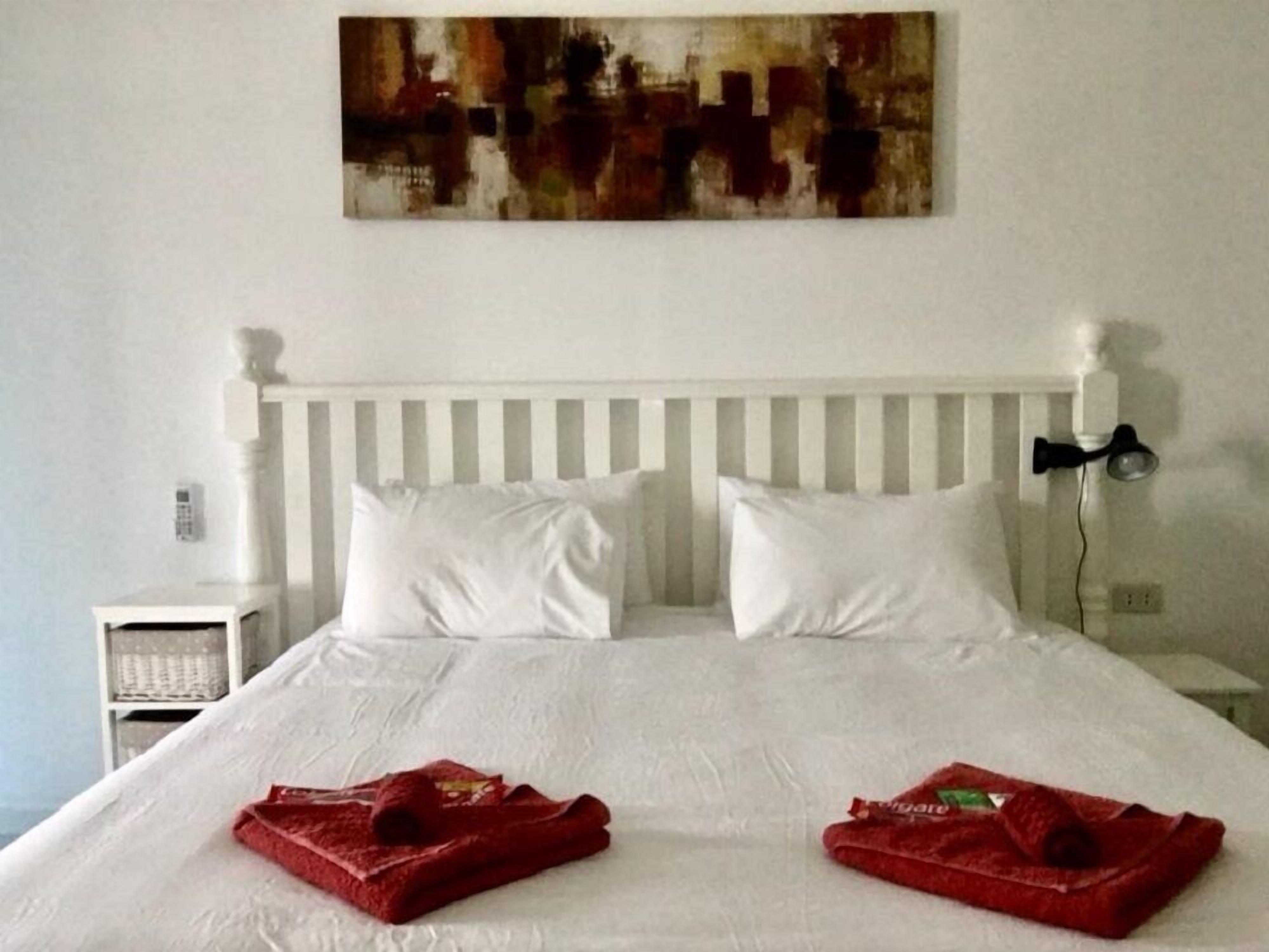 Quarto de hotel com cama branca, duas almofadas, cabeceira branca, mesa de cabeceira e uma obra de arte na parede.