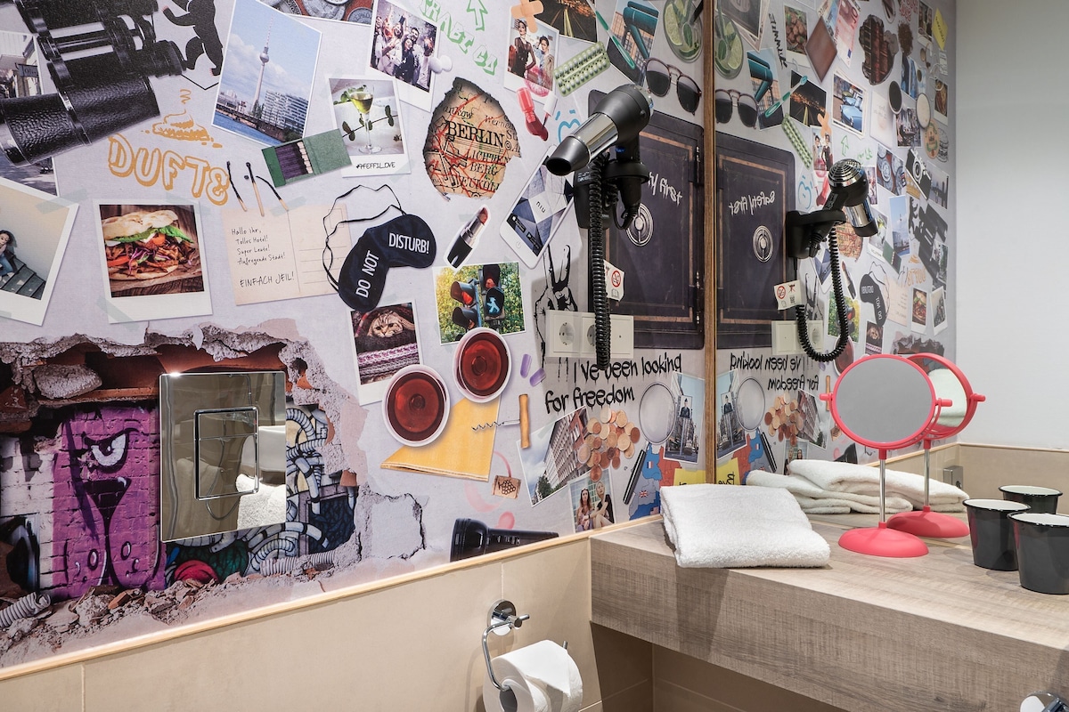 Un baño con una pared decorada con varias fotografías y carteles, un espejo y una zona de lavamanos.