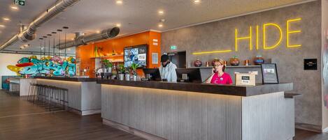 A hotel reception area with a modern design, including a reception desk, bar area, and a wall with a neon 'HIDE' sign.