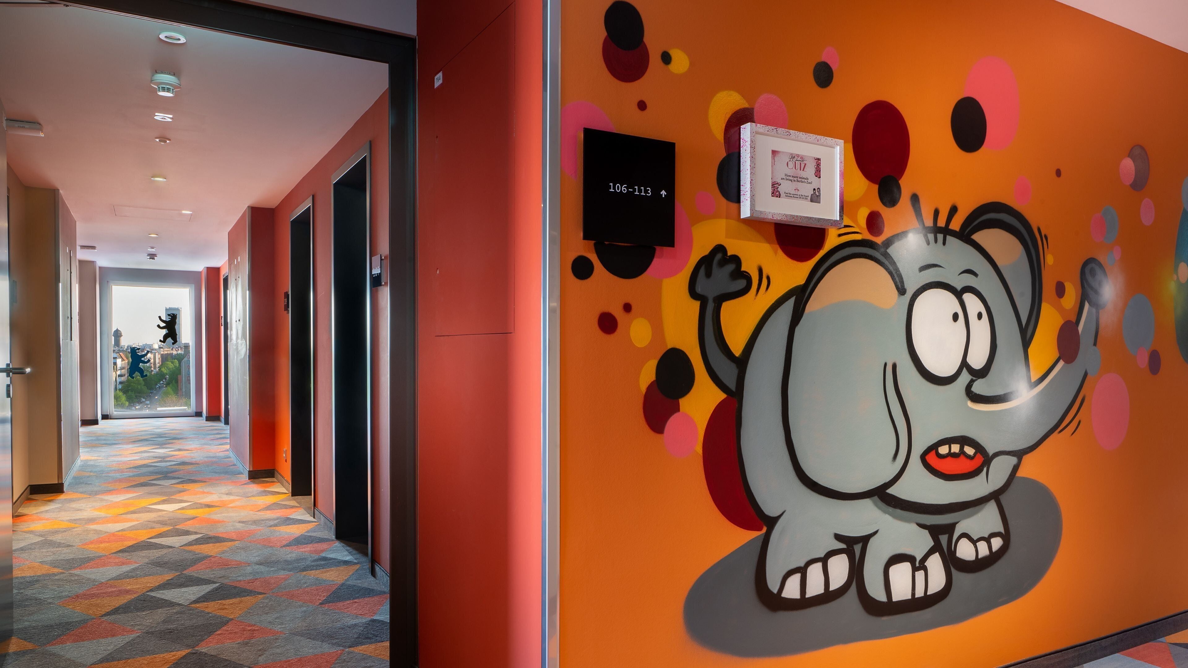 Een hotelgang met een kleurrijke muurschildering van een olifant, een bord met kamer nummers en een kamerjas patroon vloerkleed.