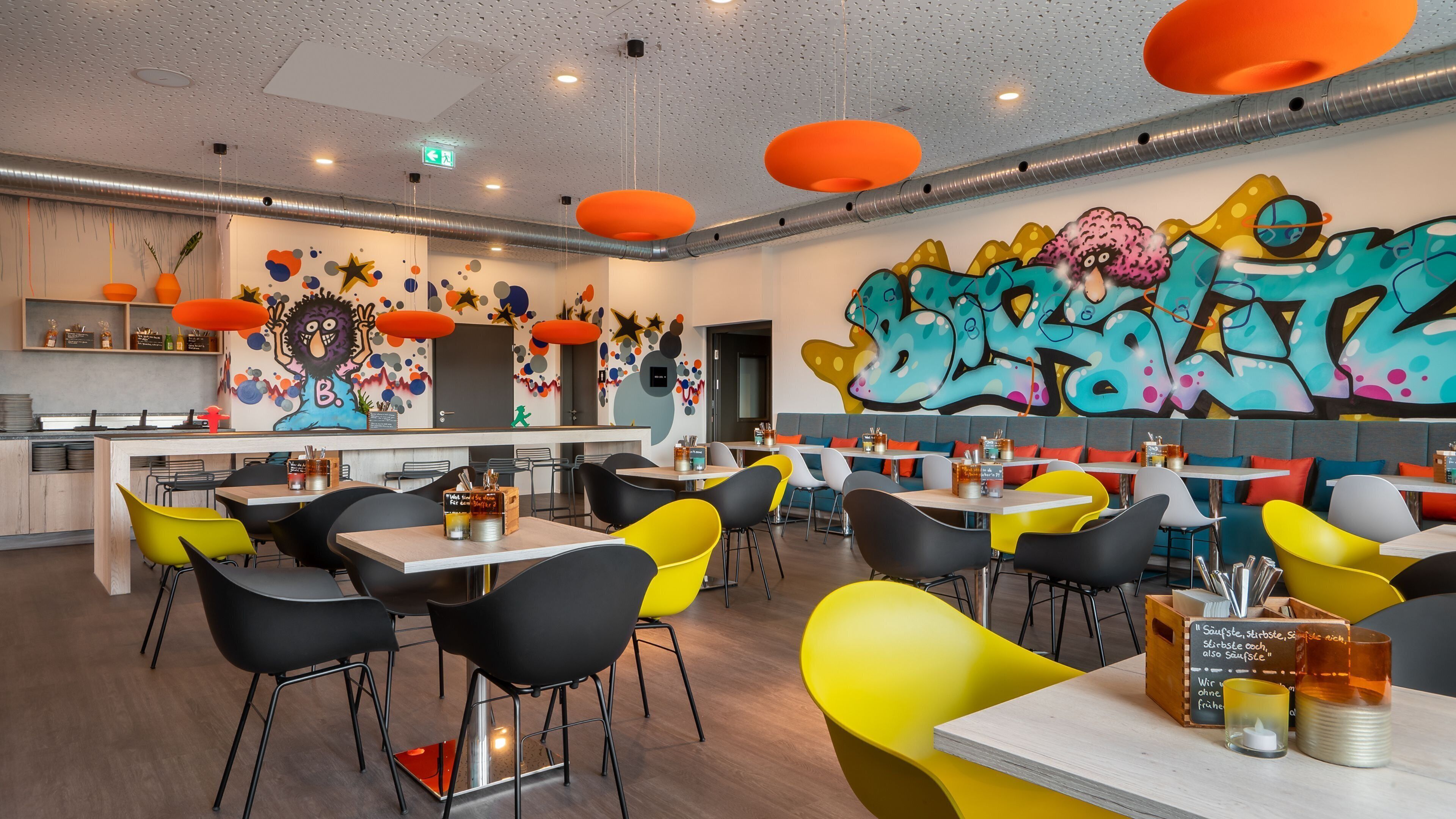Ein modernes Restaurant mit farbenfrohen Graffiti-Kunst an den Wänden, leuchtend gelb-schwarzen Stühlen und einem Holzboden.