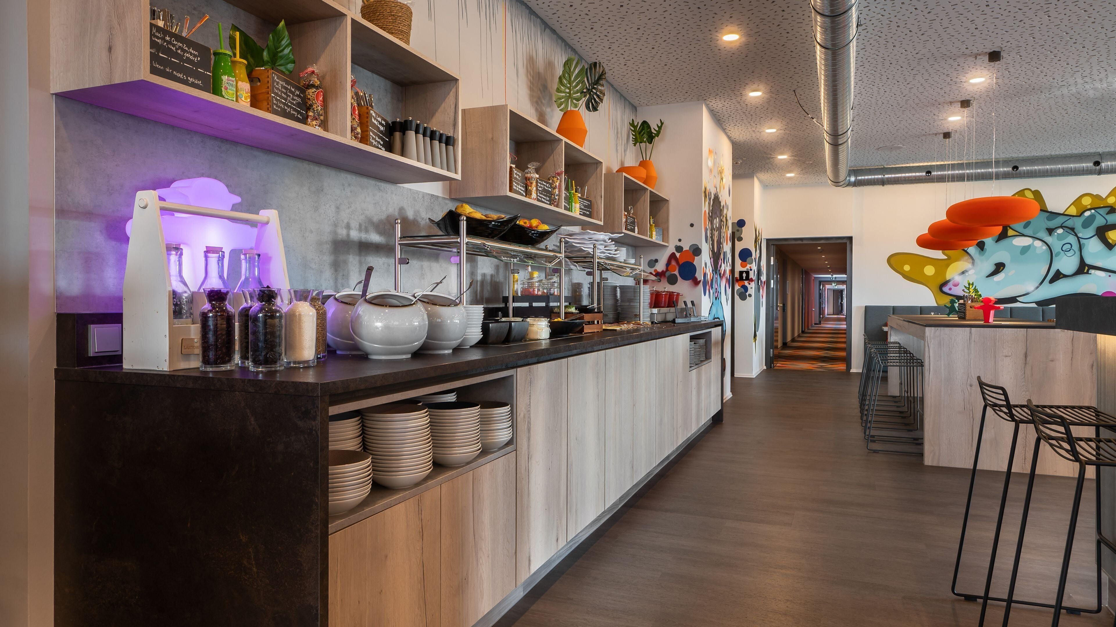 Una cocina moderna con armarios de madera, una encimera larga con distintos utensilios de cocina y un mural colorido en la pared.