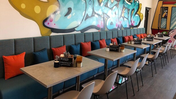A restaurant with long tables and booths, a colorful graffiti wall, and a modern ceiling design. - Holiday Inn - the niu, Hide Berlin Friedrichshain by IHG (Berlin)