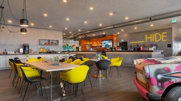 Un restaurant moderne avec des chaises jaunes et noires, un coin bar et un panneau néon sur lequel on peut lire « HIDE ».