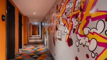 A hotel corridor with a colorful geometric patterned floor, a wall with cartoon character graffiti, and doors marked with room numbers.