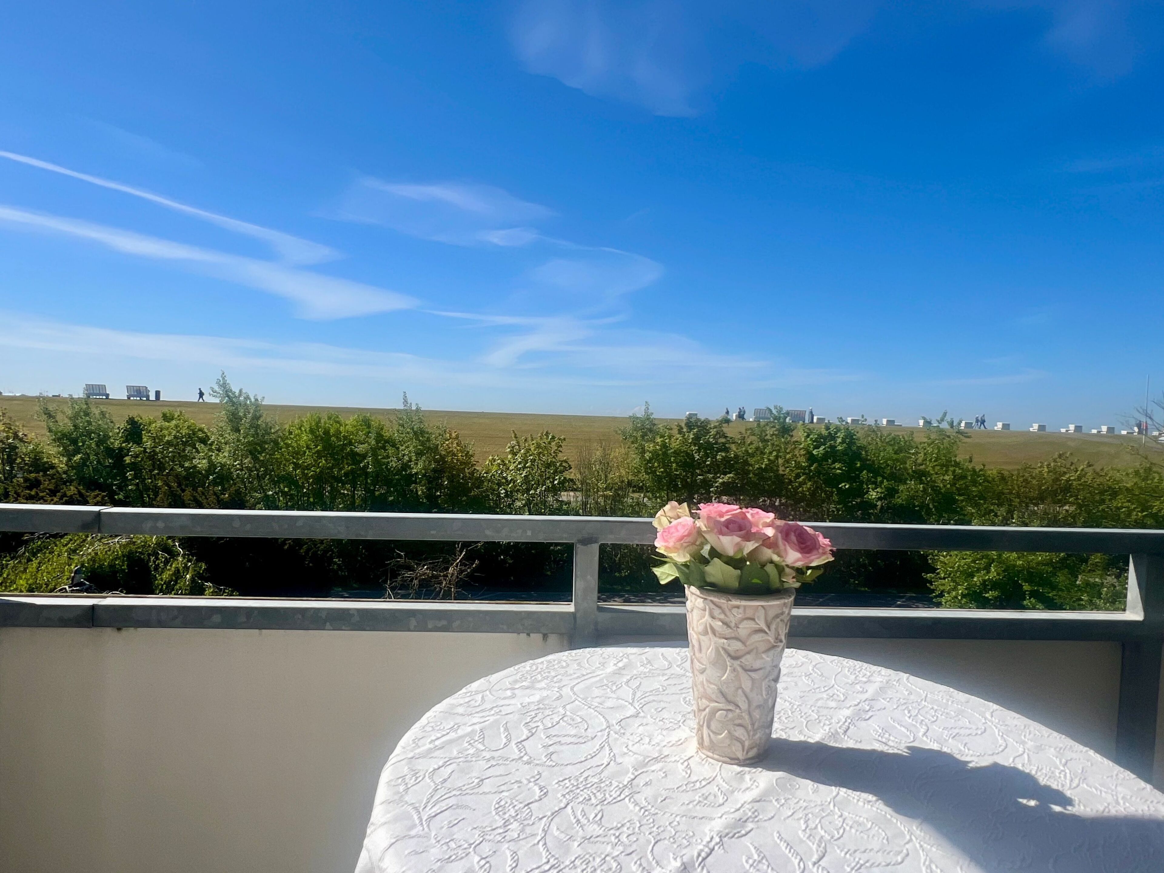 Un balcon avec une table blanche et un vase de fleurs, d’où l’on peut admirer un paysage verdoyant et des bâtiments situés au loin.