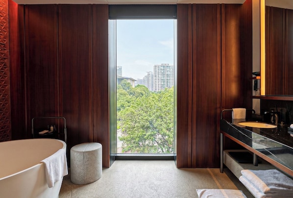 A modern bathroom with a bathtub, a round stool, a vanity with a mirror, and a view of greenery and buildings through a large window. - Lohkah Hotel & Spa (Xiamen)