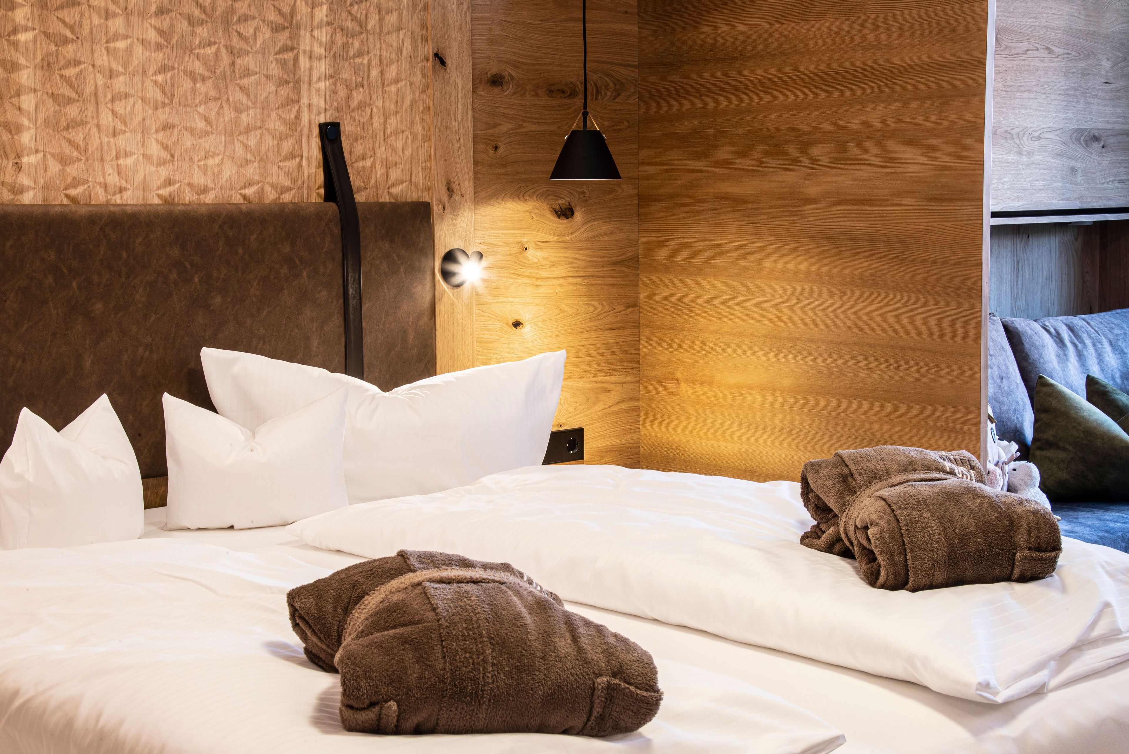 A neatly made bed with white linens and brown towels, a black pendant light, and a wooden wall with a geometric pattern.