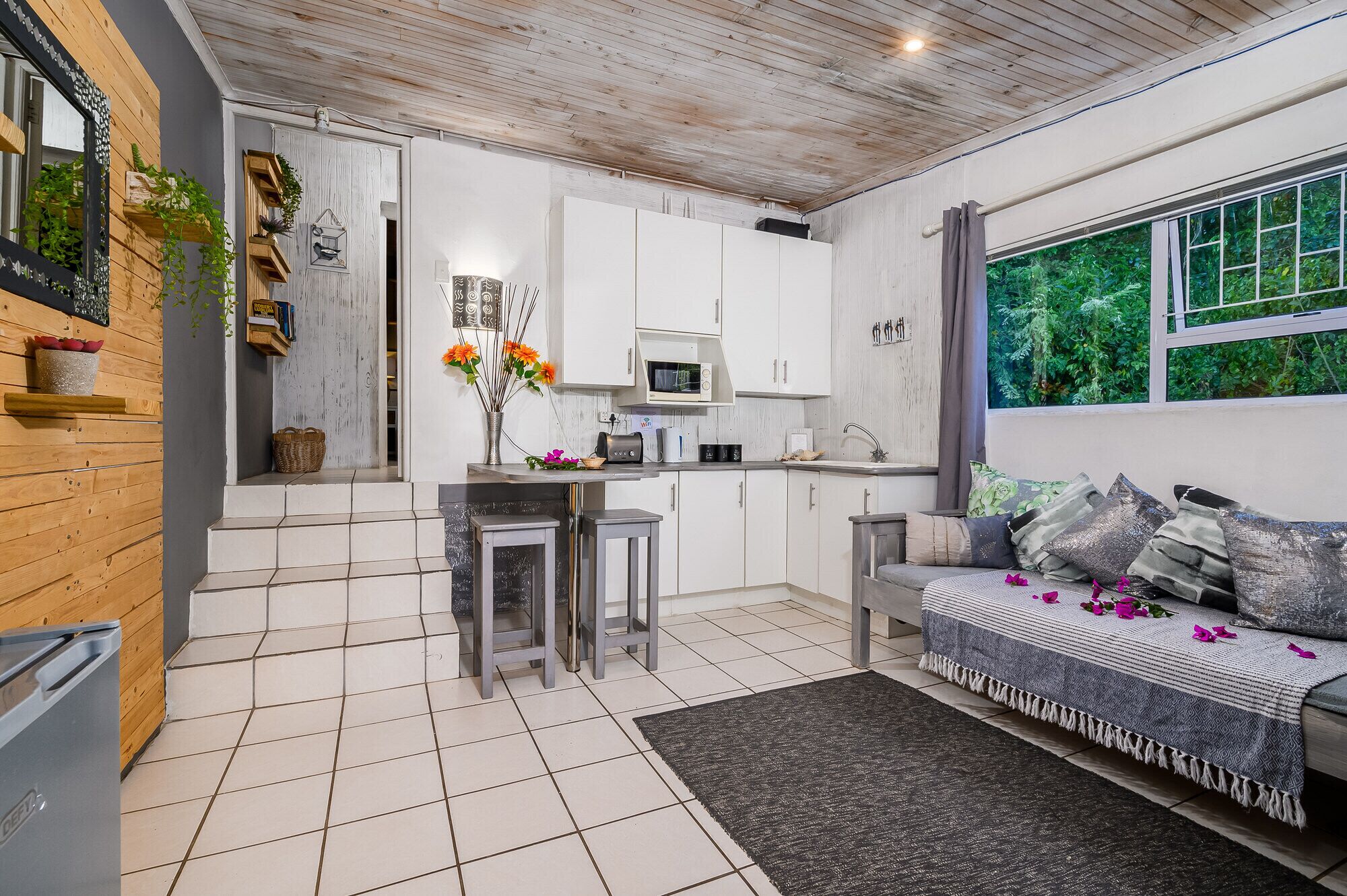 A modern kitchen with white cabinets, a microwave, and a sink. There is a dining area with a table and chairs, and a sofa with a patterned throw.