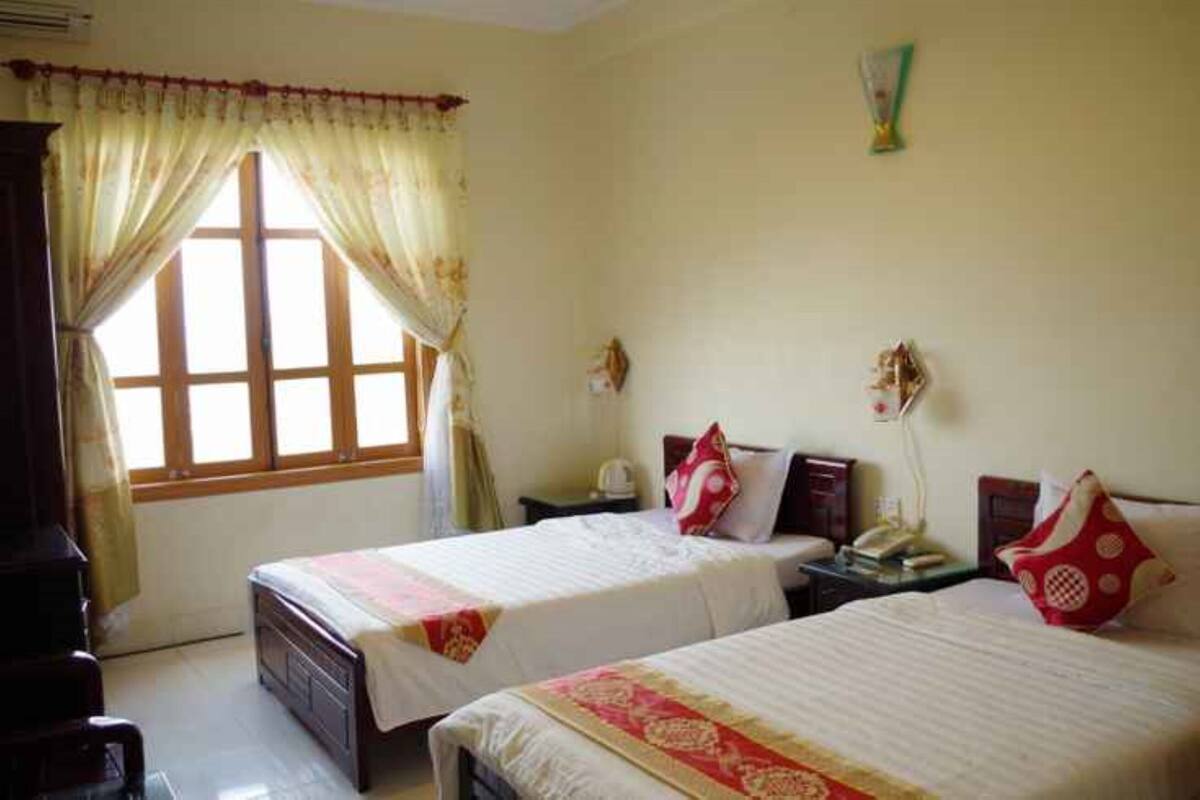 A hotel room with two beds, a window with curtains, a bedside table with a lamp, and a wall-mounted telephone.