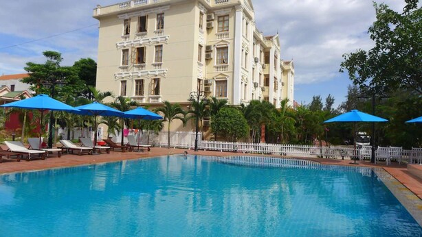 Una piscina con sillas de descanso y sombrillas, rodeada por una cerca blanca y palmeras, con un edificio grande al fondo.