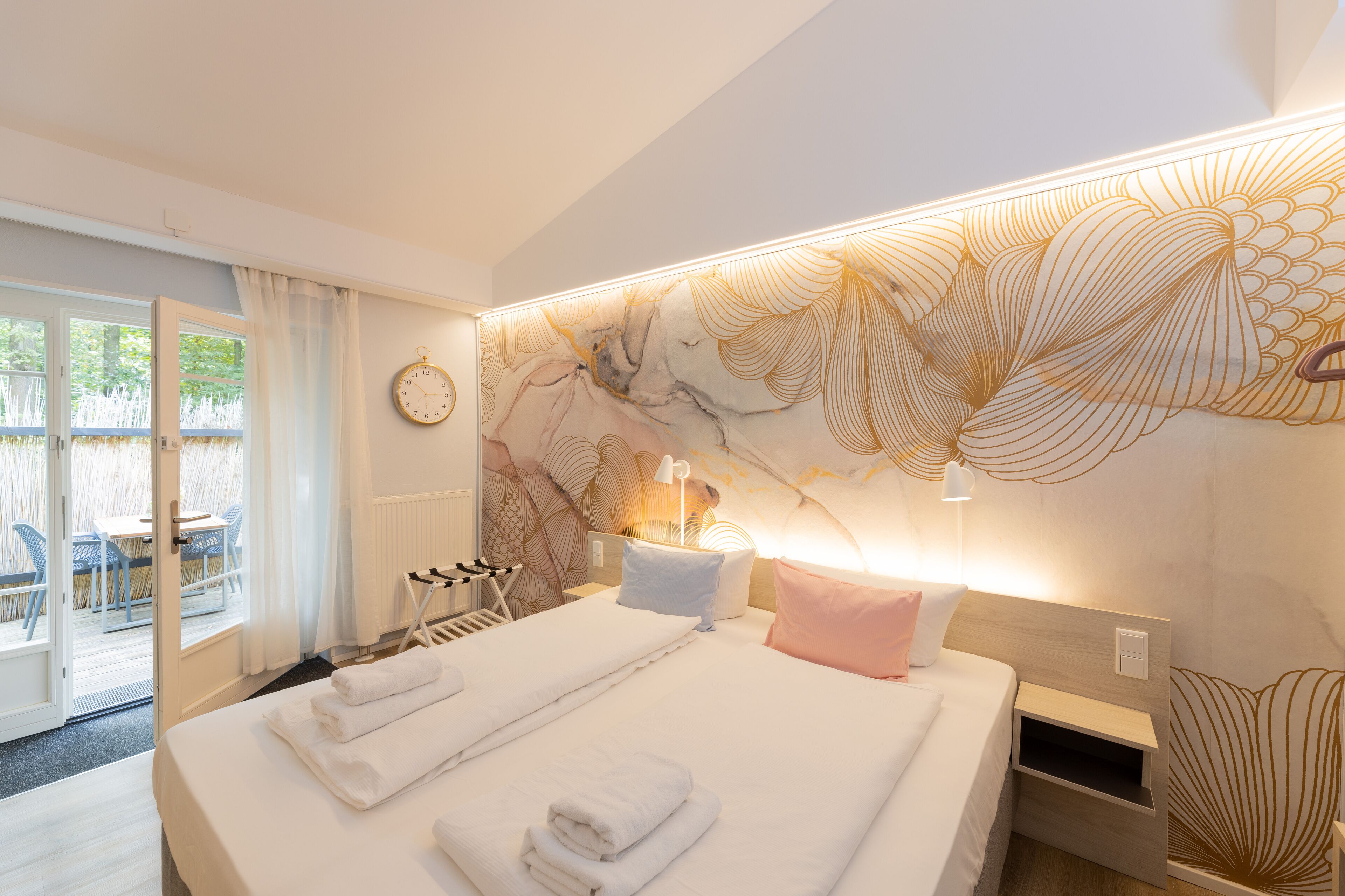 Ein Hotelzimmer mit einem großen Bett, einem Nachttisch, einer Wanduhr und Blick ins Freie.