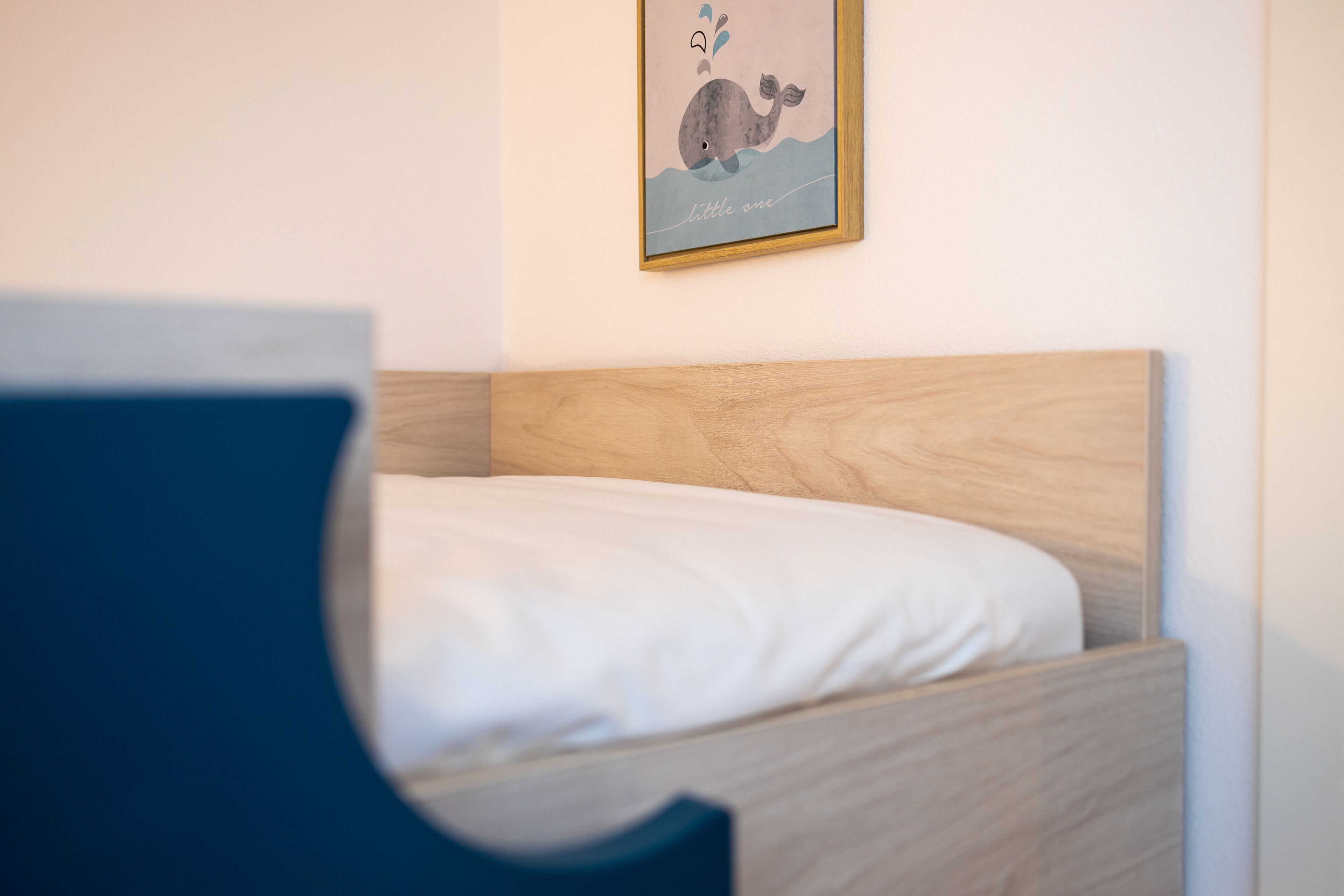 A neatly made bed with a wooden headboard, a wall-mounted picture of a whale, and a blue chair in the foreground.