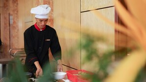 A chef in a black uniform with a white chef's hat, preparing food in a kitchen with wooden cabinets and a red basket of ingredients. - SeaStar Hotel Ha Long (Bai Chay)