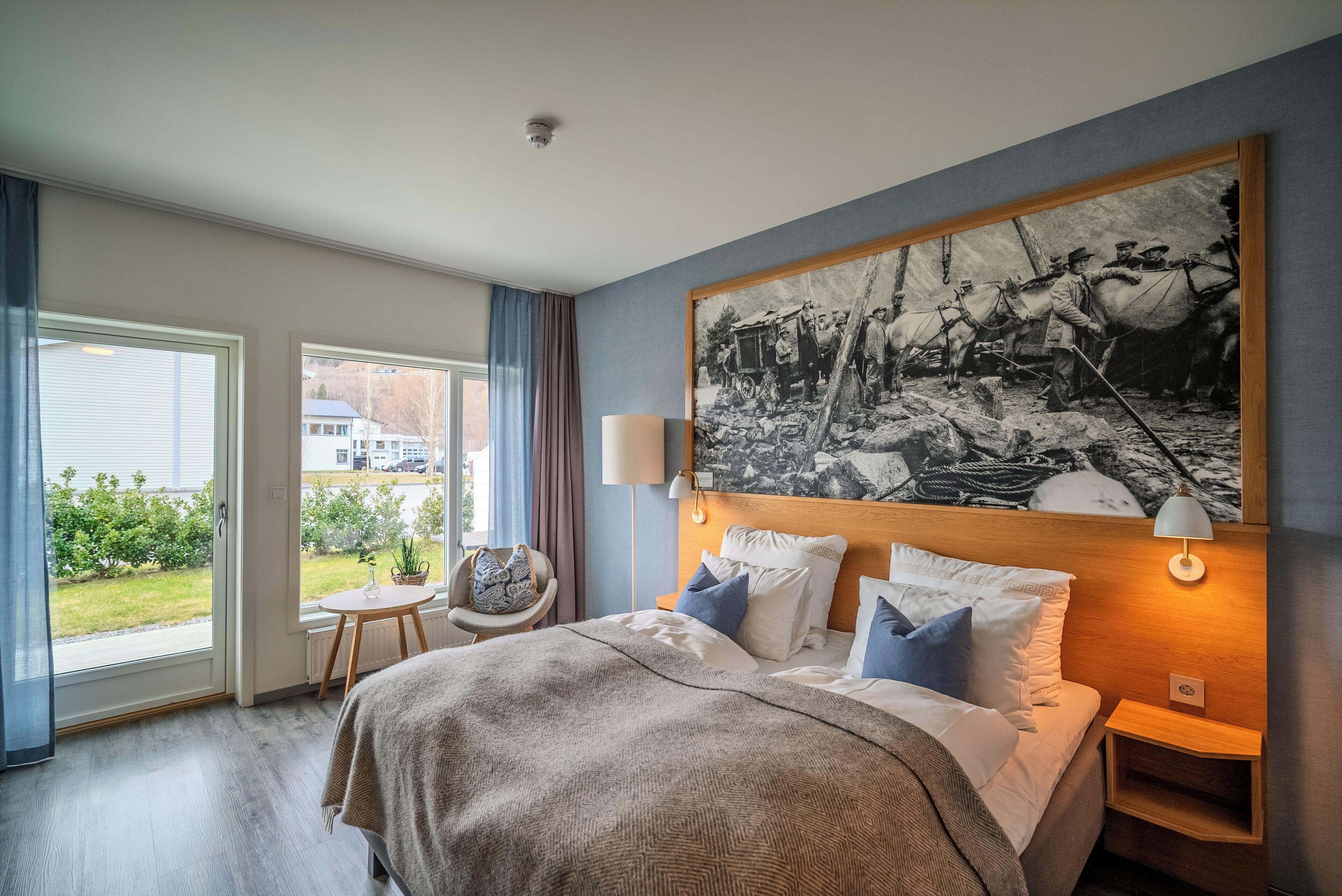 A hotel room with a large bed, a window with a view of the outdoors, a painting on the wall, and a small table with a chair.