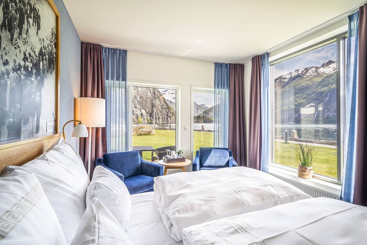 Ein Hotelzimmer mit einem Bett, zwei Stühlen, einem kleinen Tisch und Blick auf Berge und See durch große Fenster.