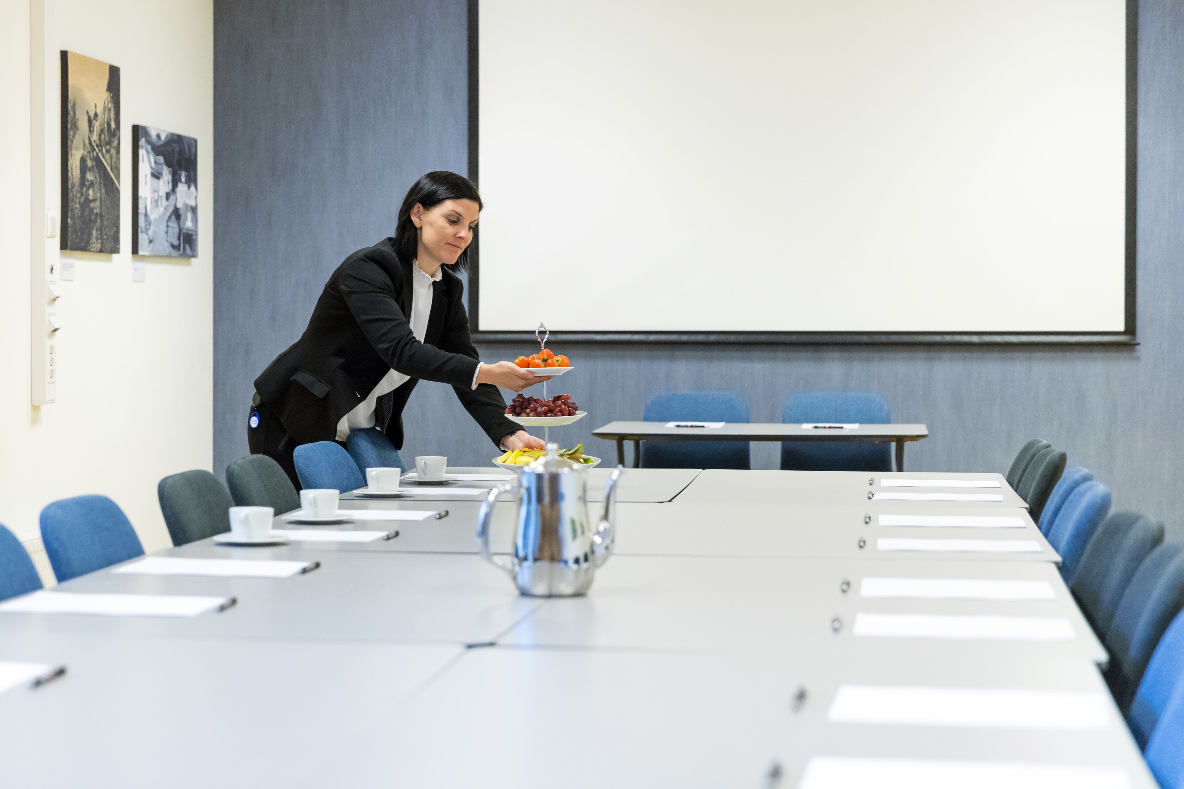 Une salle de conférence avec une longue table, des chaises, un écran de projection et une personne qui sert à manger.