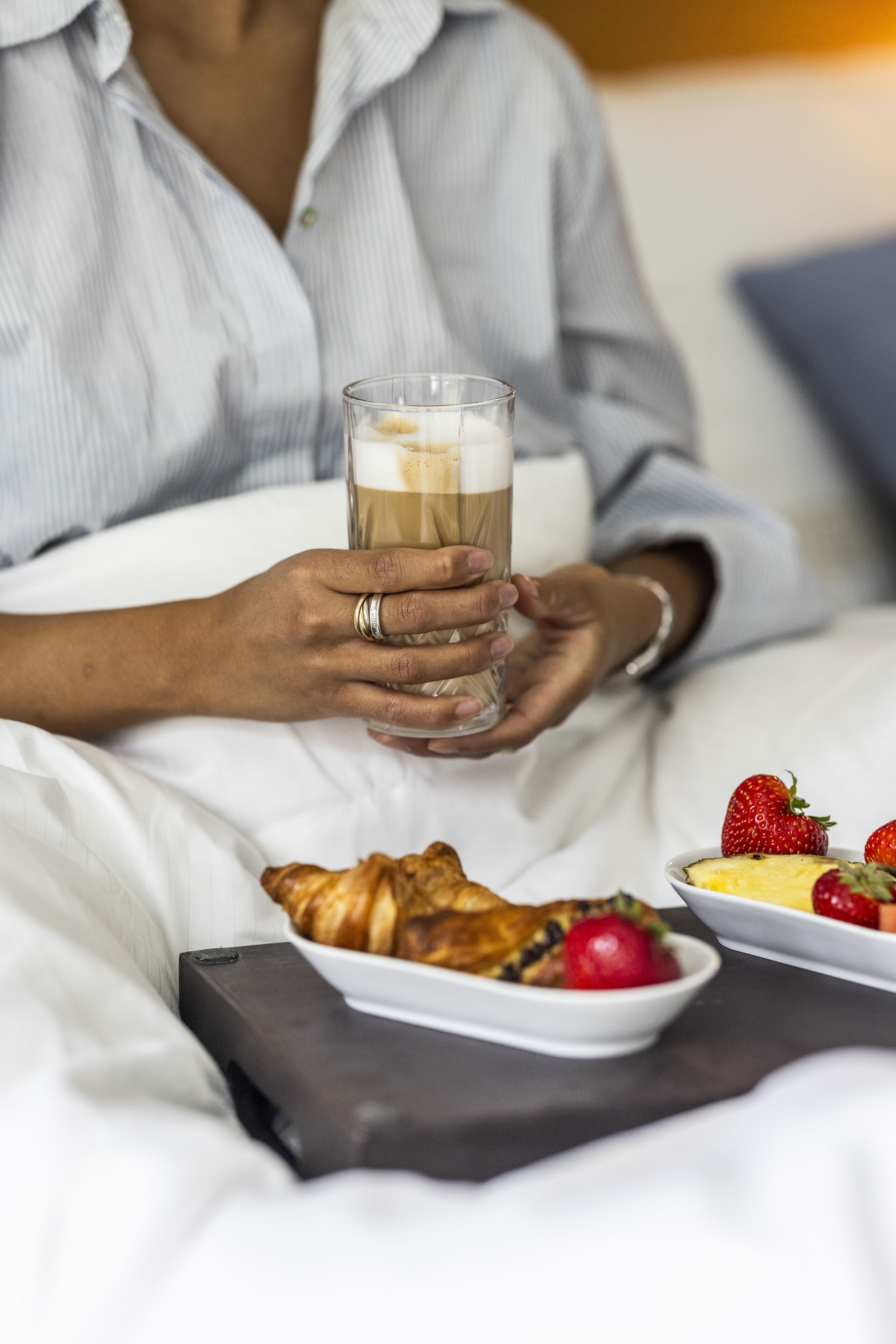 A person holding a glass of coffee, a plate of croissants, and a bowl of fruit on a bed.