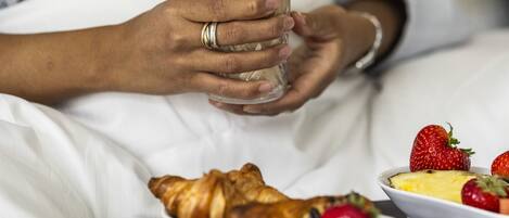 A person holding a glass of coffee, a plate of croissants, and a bowl of fruit on a bed.