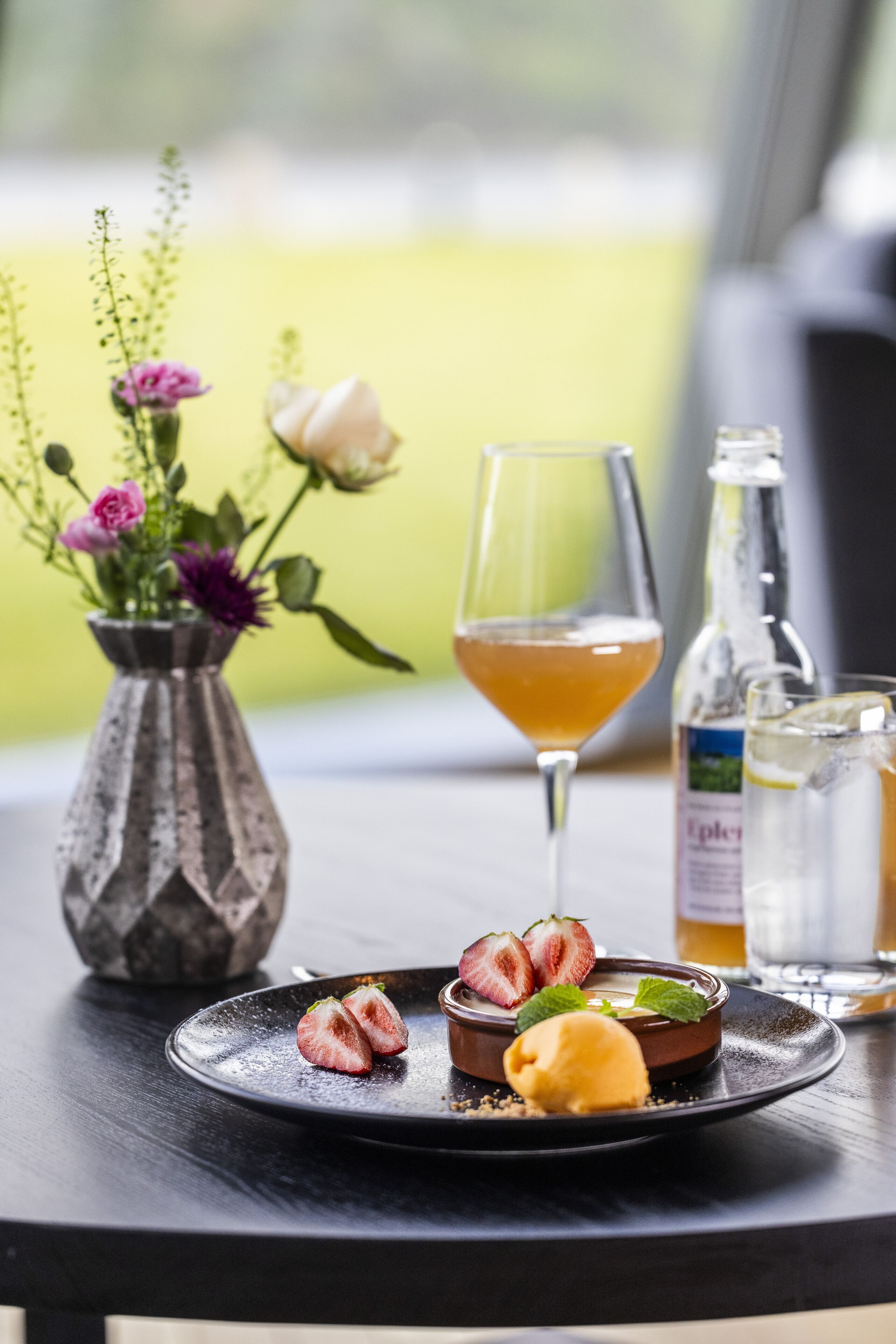 Un plato con fresas, una bola de helado, un vaso de bebida color ámbar, una botella de agua y un florero con flores sobre una mesa.