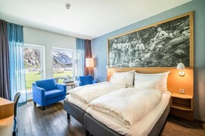 A hotel room with a large bed, a blue armchair, a desk, and a window with curtains. - Valldal Fjordhotell - by Classic Norway Hotels (Fjord)