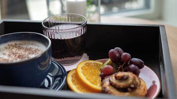 Una bandeja con una taza de café, un vaso de bebida oscura, un plato con un rollo de canela, uvas y una rodaja de naranja.