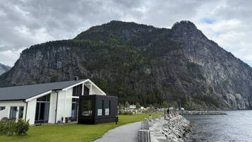 A modern house with a white exterior and a black extension, situated by a rocky waterfront with a mountainous backdrop.