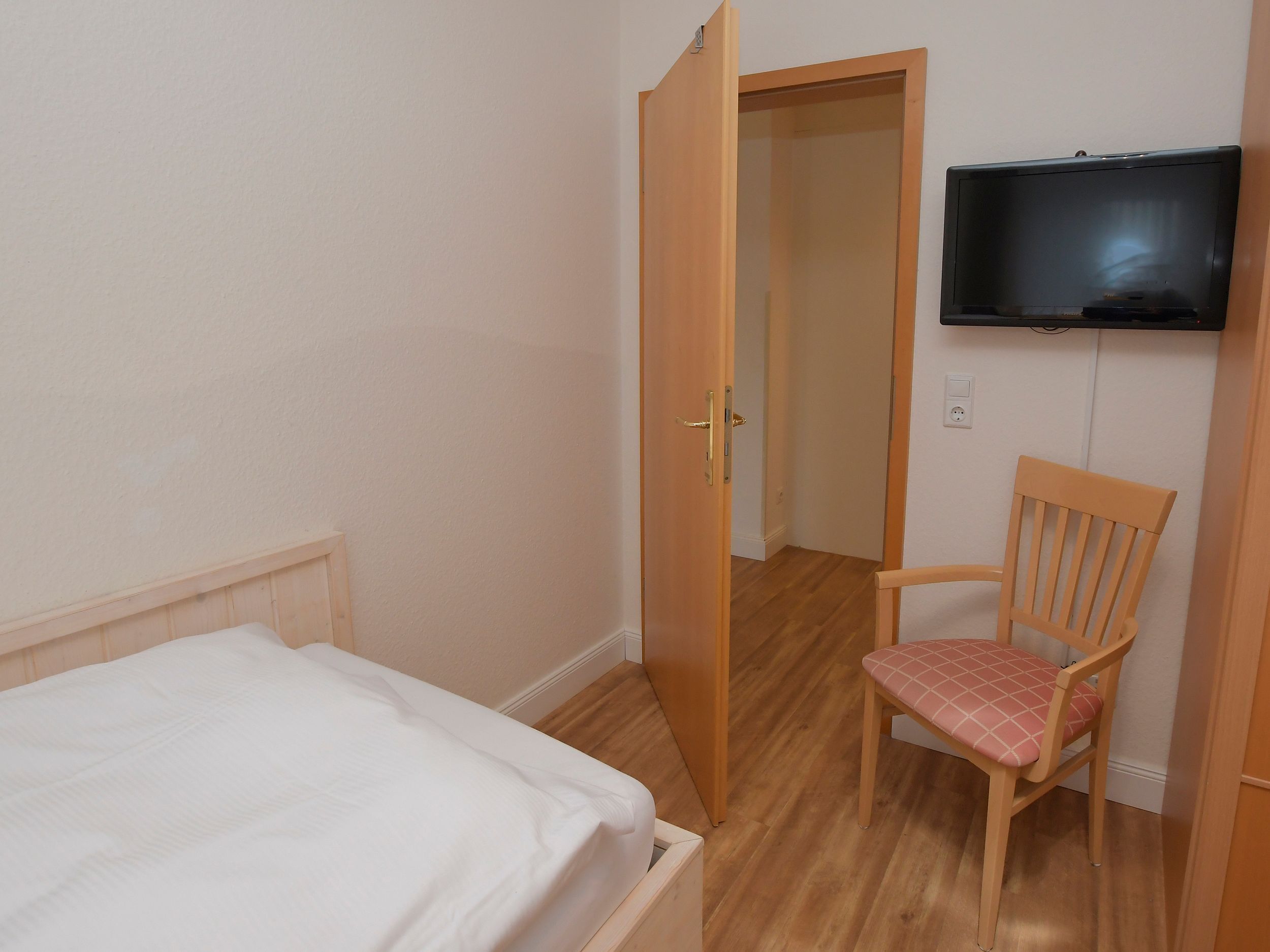 Une chambre à coucher comprenant un lit, une chaise en bois, un téléviseur fixé au mur et une porte donnant sur une autre pièce.