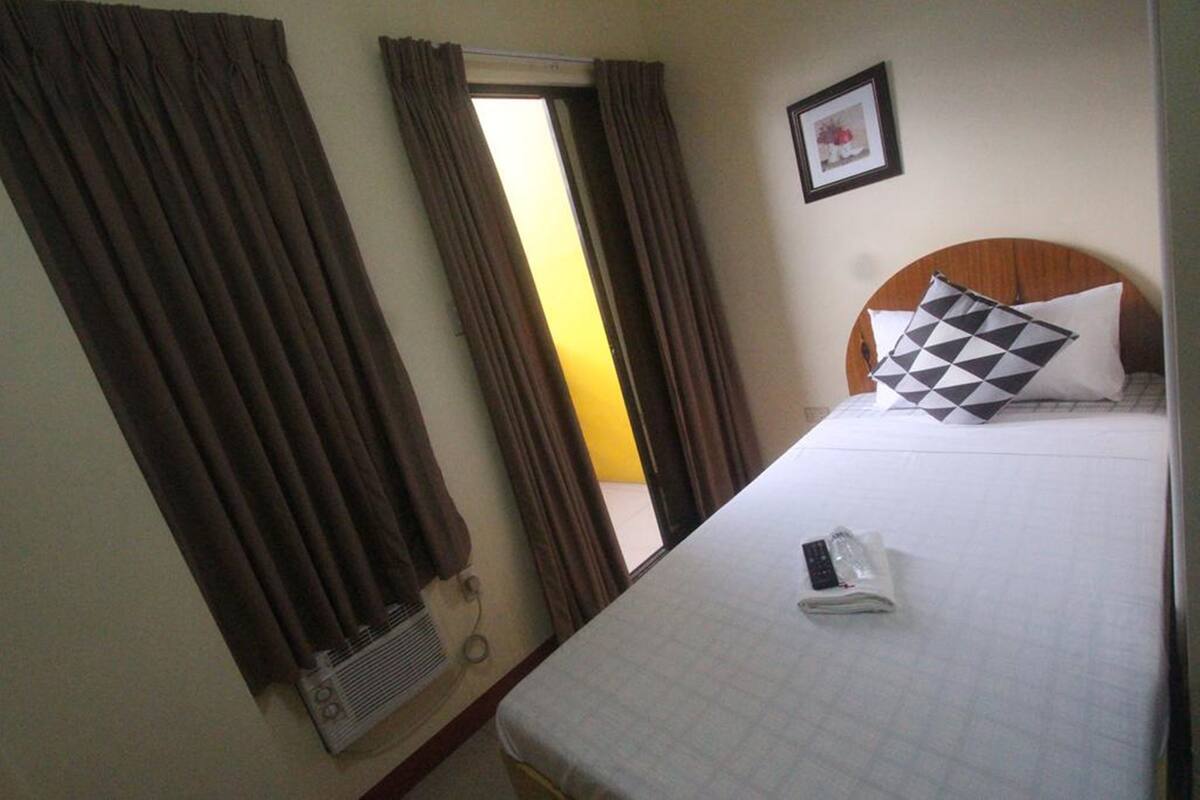 A hotel room with a bed, curtains, a framed picture on the wall, and a window with curtains partially drawn.
