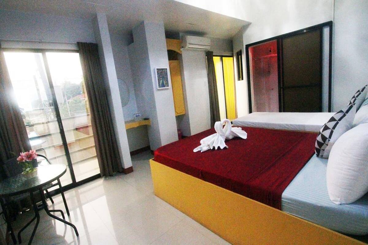 A hotel room with a bed, a small table with a chair, a balcony with curtains, and a window.