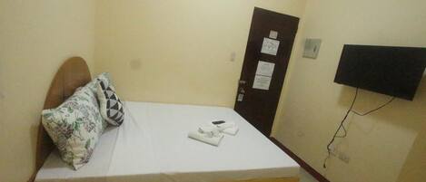 Una habitación pequeña con cama individual, cabecera de madera, colchón blanco y almohada estampada. Se observa una puerta con un aviso y un televisor montado en la pared.