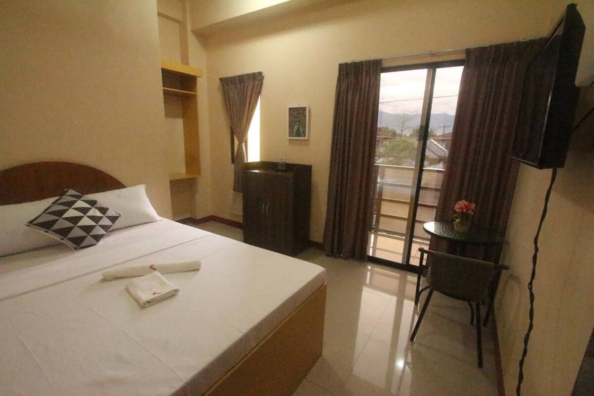 A hotel room with a bed, a desk, a chair, a TV, and a view of the outdoors.