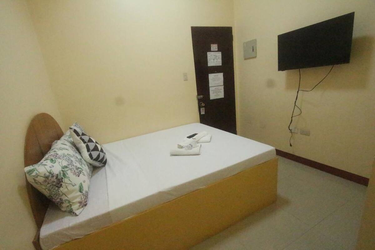 A small, single-bed room with a wooden headboard, a white mattress, and a floral pillow. There is a door with a bulletin board and a flat-screen TV mounted on the wall.