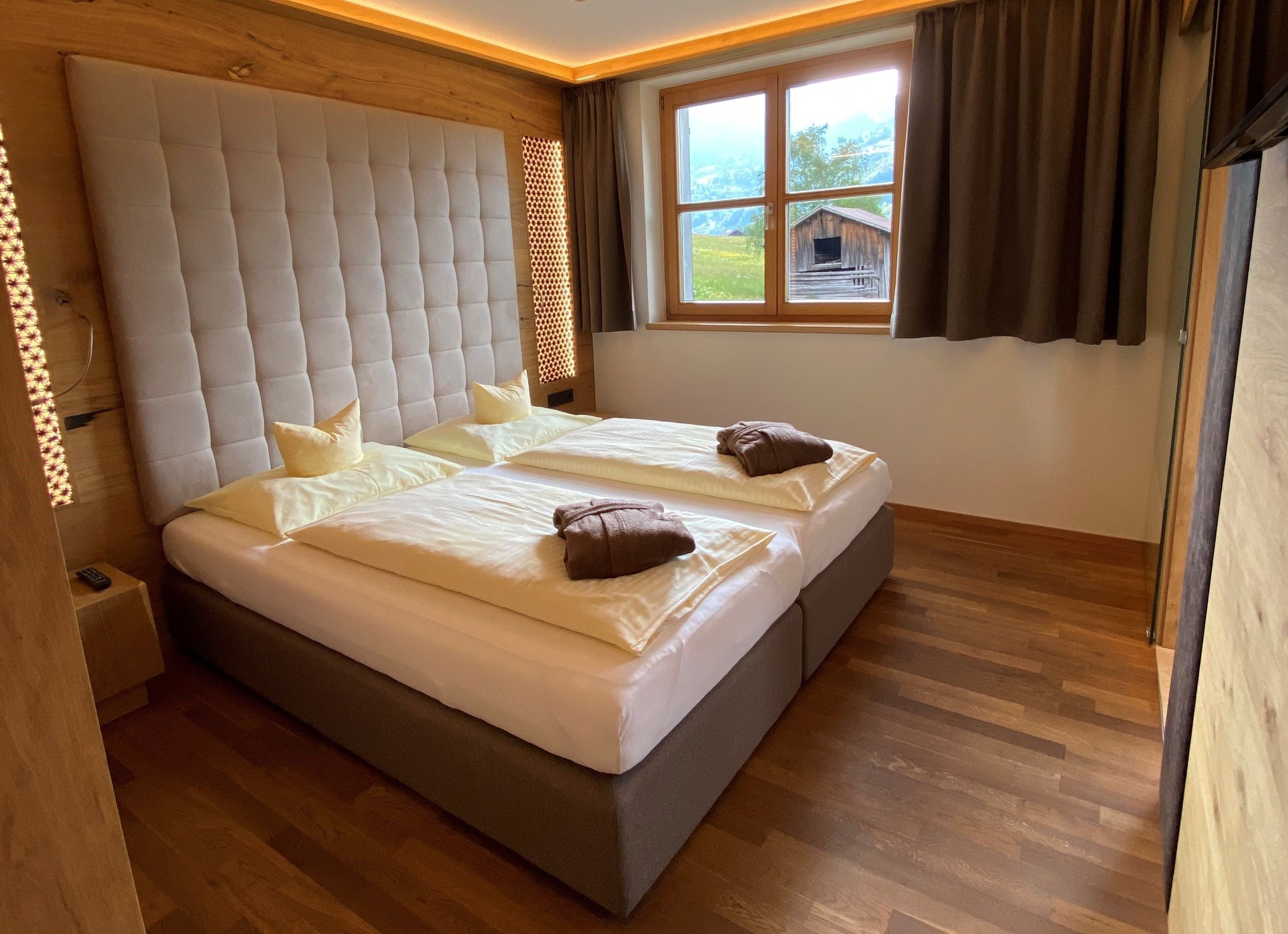 A double bed with a tufted headboard, two pillows, and a window with a view of a wooden cabin.