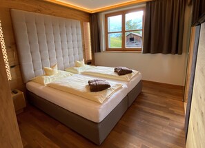 Een tweepersoonsbed met een gestuukt hoofdbord, twee kussens, en een raam met uitzicht op een houten hut.