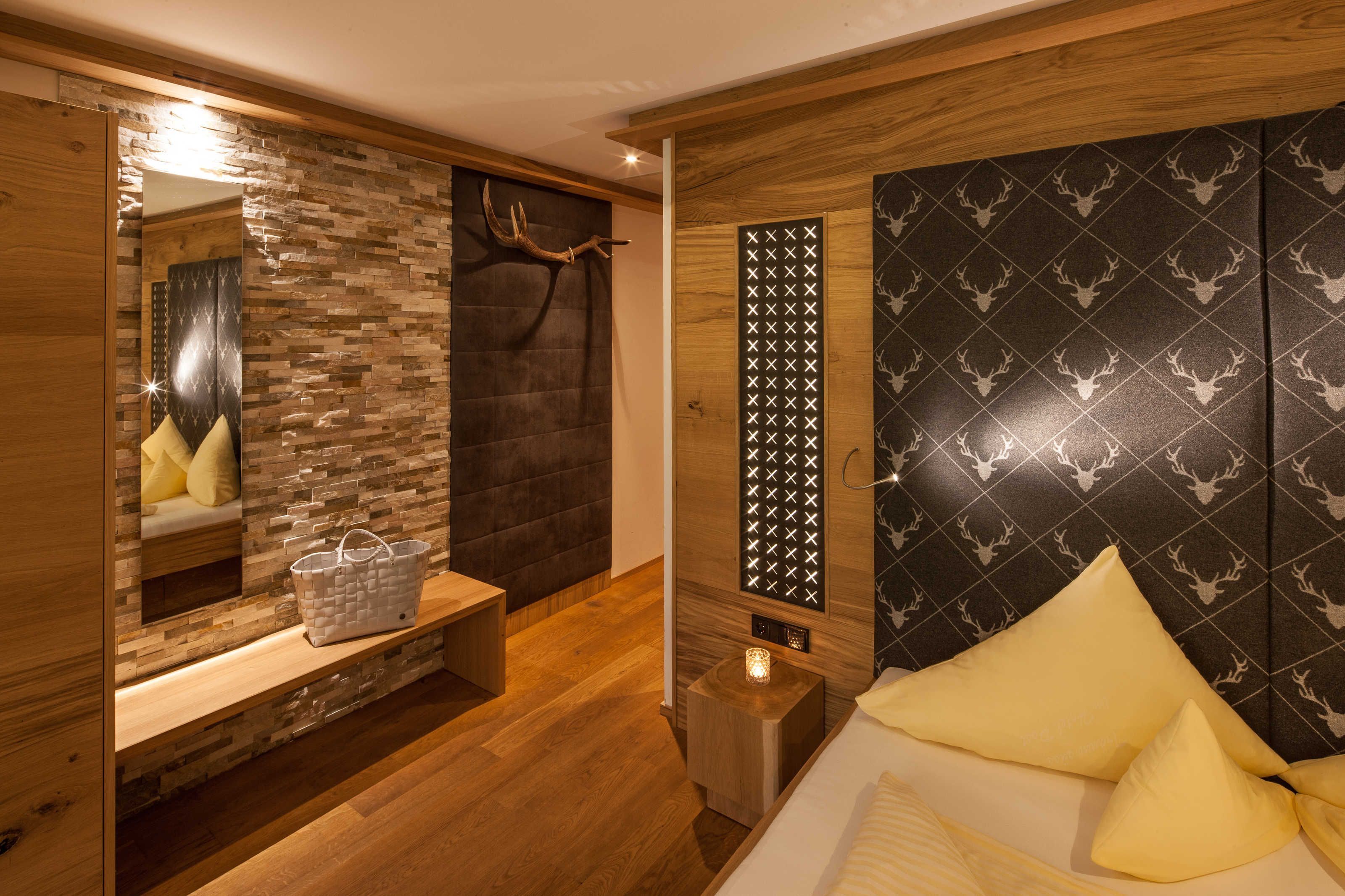 Een moderne kamer met een houten vloer, een bed met gele kussens, een bankje met een mand, een spiegel en een muur met een hertenpatroon.