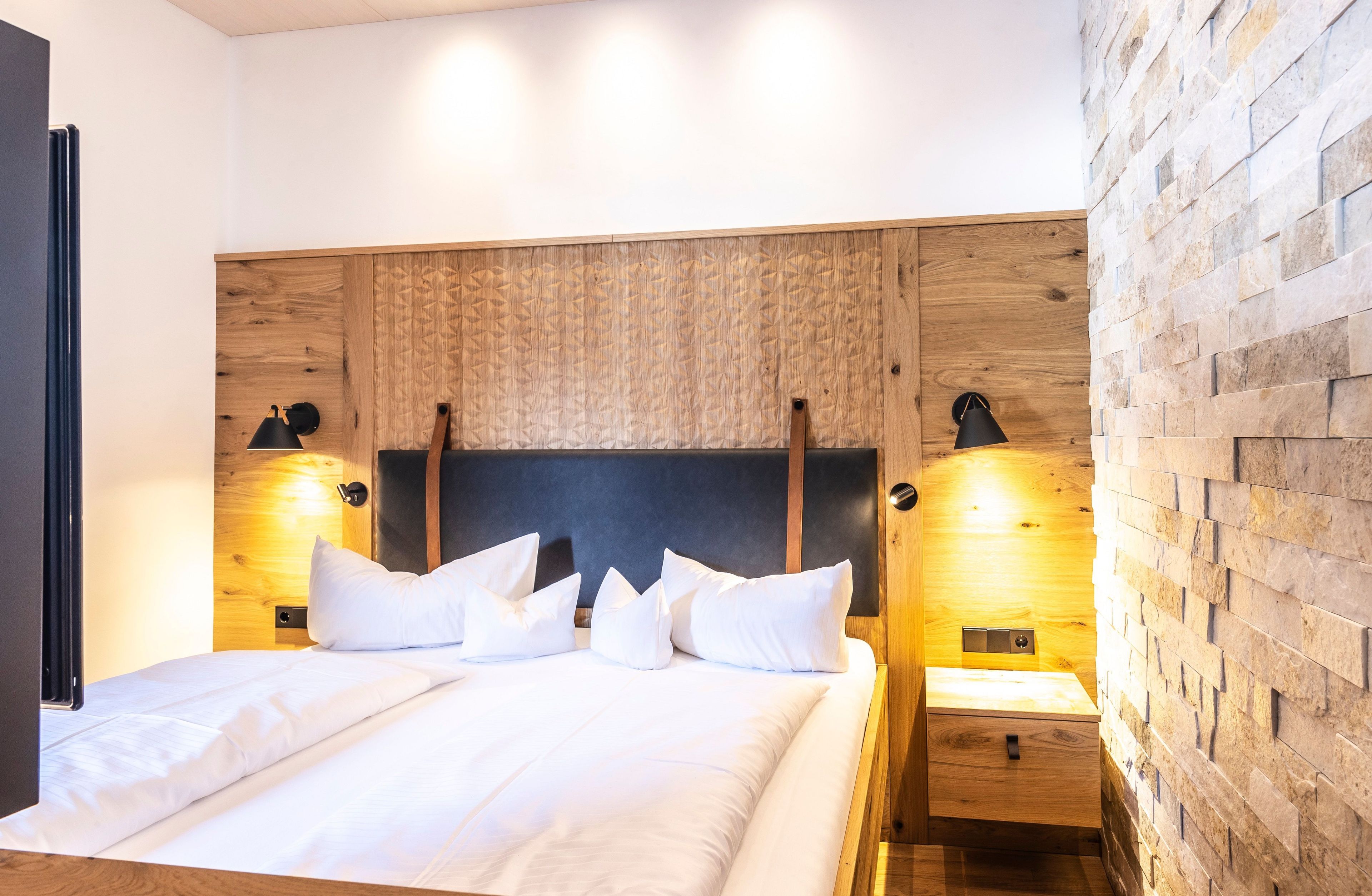 A modern bedroom with a wooden headboard, a bed with white linens, two bedside tables with lamps, and a brick-patterned wall.