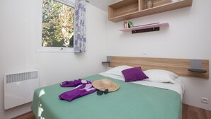 A bedroom with a bed, a window with a view of trees, a radiator, and shelves with decorative items. - Camping le Paradis (Talmont-Saint-Hilaire)