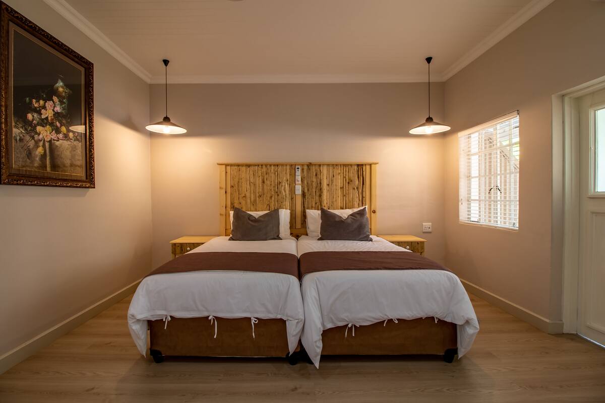 A bedroom with two beds, wooden headboard, pendant lights, a painting, and a window with blinds.
