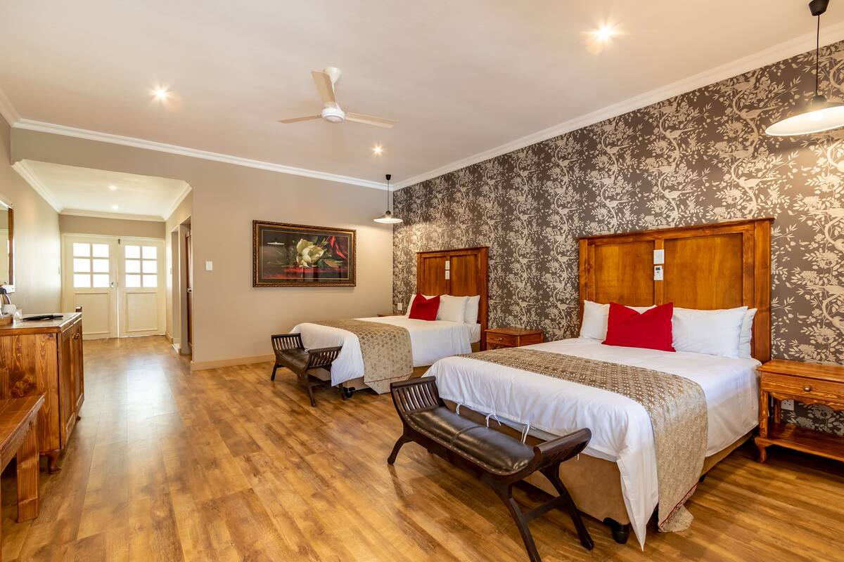 A hotel room with two beds, wooden headboards, a bench, a desk, and a ceiling fan.