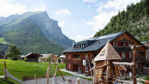Un terrain de jeux avec une structure d’escalade en bois et un toboggan, le tout situé devant un paysage de montagnes et un bâtiment au toit rouge.